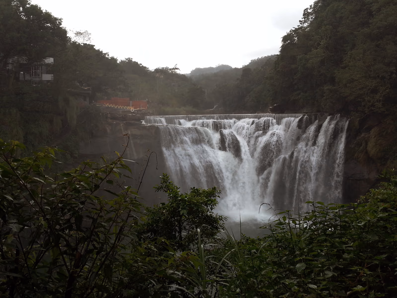 A waterfall in a lush forest with a traditional Chinese-style tower in the background.